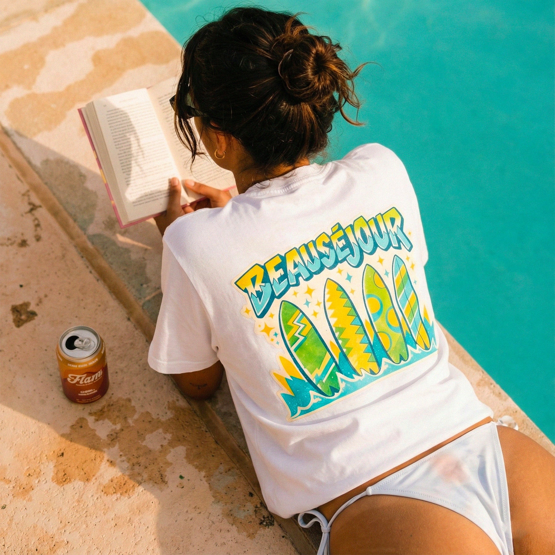 Femme au bord d'une piscine a bali en train de lire. elle porte un tshirt beauséjour lifestyle imprimé planche de surf dans le dos.