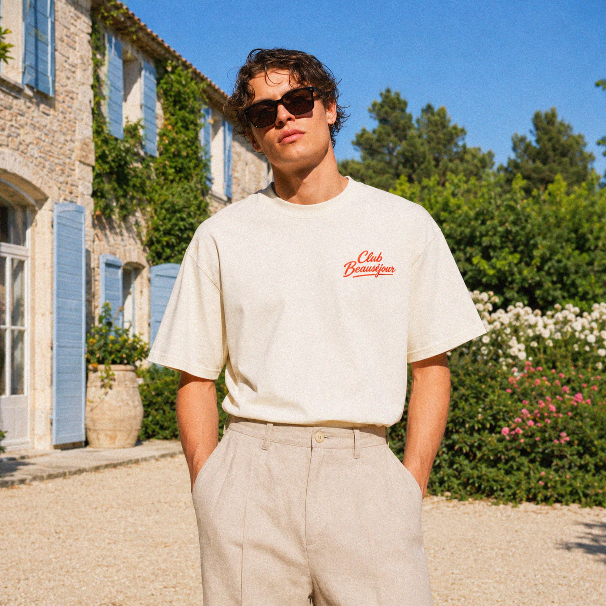 T-shirt boxy crème Club Beauséjour avec logo rouge vintage sur la poitrine, porté dans une maison du Sud inspiration Riviera.