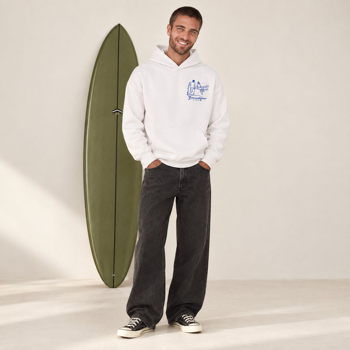 Hoodie Biarritz after surf blanc porté par un homme souriant, debout à côté d'une planche de surf verte
