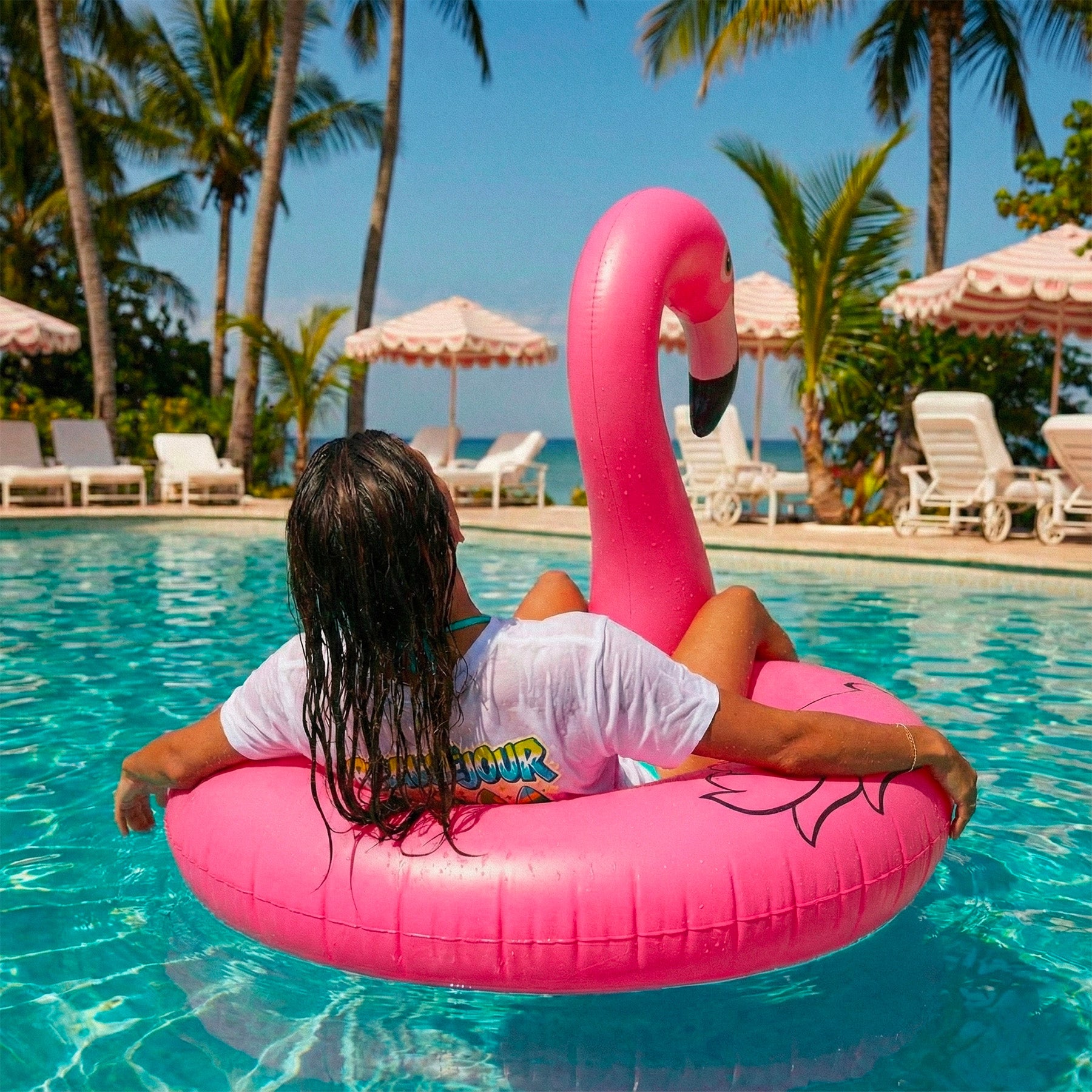 Une personne vue de dos se détend sur une bouée flamant rose dans une piscine turquoise, entourée de palmiers et de transats, avec la mer visible à l’arrière-plan sous un ciel ensoleillé. Le model porte un tshirt lifestyle femme beausejour france. Planche de sufrf sur le tshirt en coton bio.