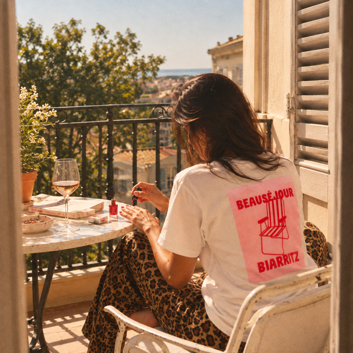 T-shirt blanc Beauséjour Biarritz avec grand imprimé rouge et rose chaise de plage au dos, porté par une femme sur un balcon ensoleillé face à la mer.