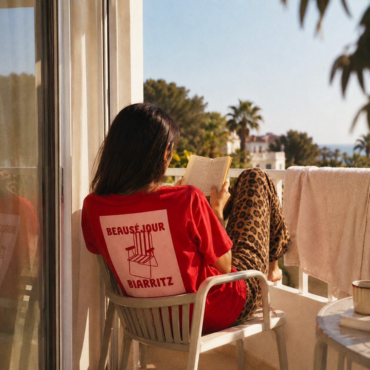 T-shirt rouge Beauséjour Biarritz avec grand imprimé chaise rose et rouge au dos, porté par une femme sur un balcon ensoleillé face à la mer, vue 3.