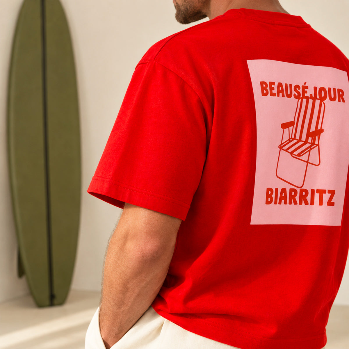 T-shirt rouge Beauséjour Biarritz porté par un homme souriant, debout à côté d'une planche de surf verte