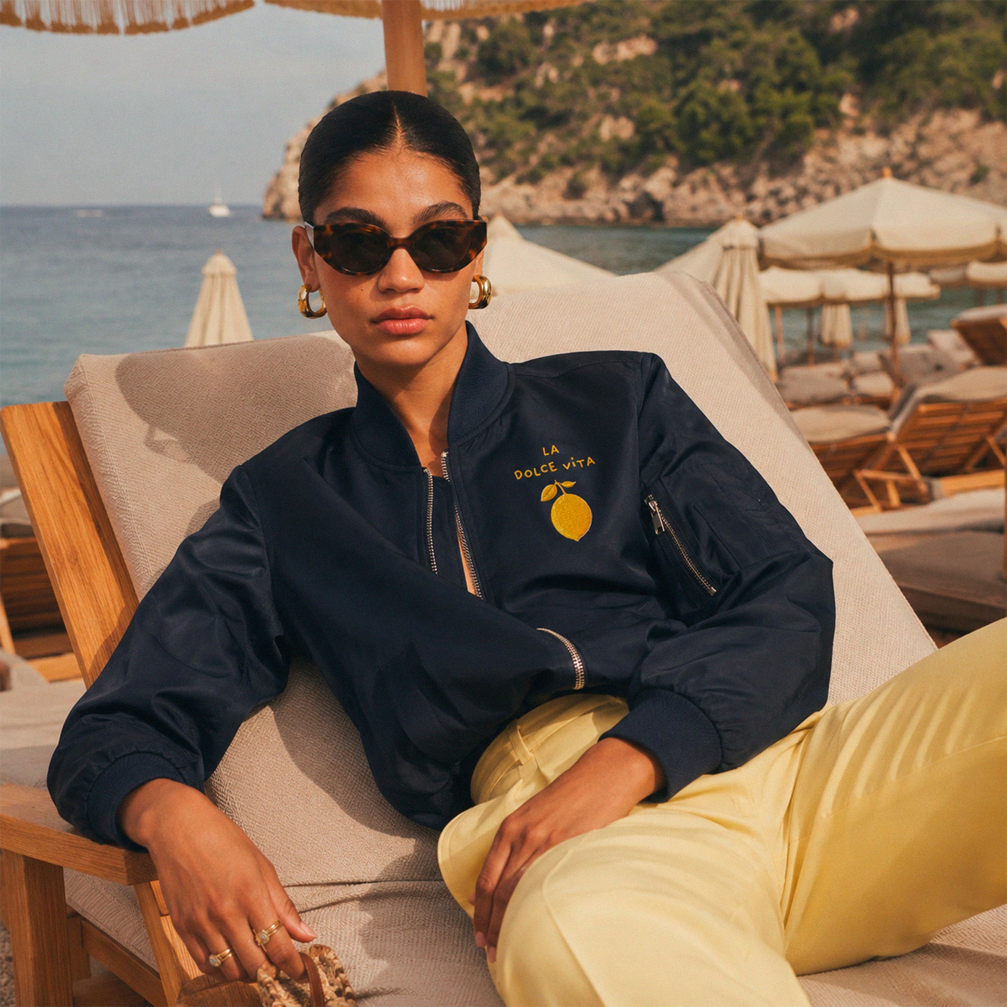 Femme portant un bomber bleu marine Dolce Vita citron sur une plage mediterraneenne