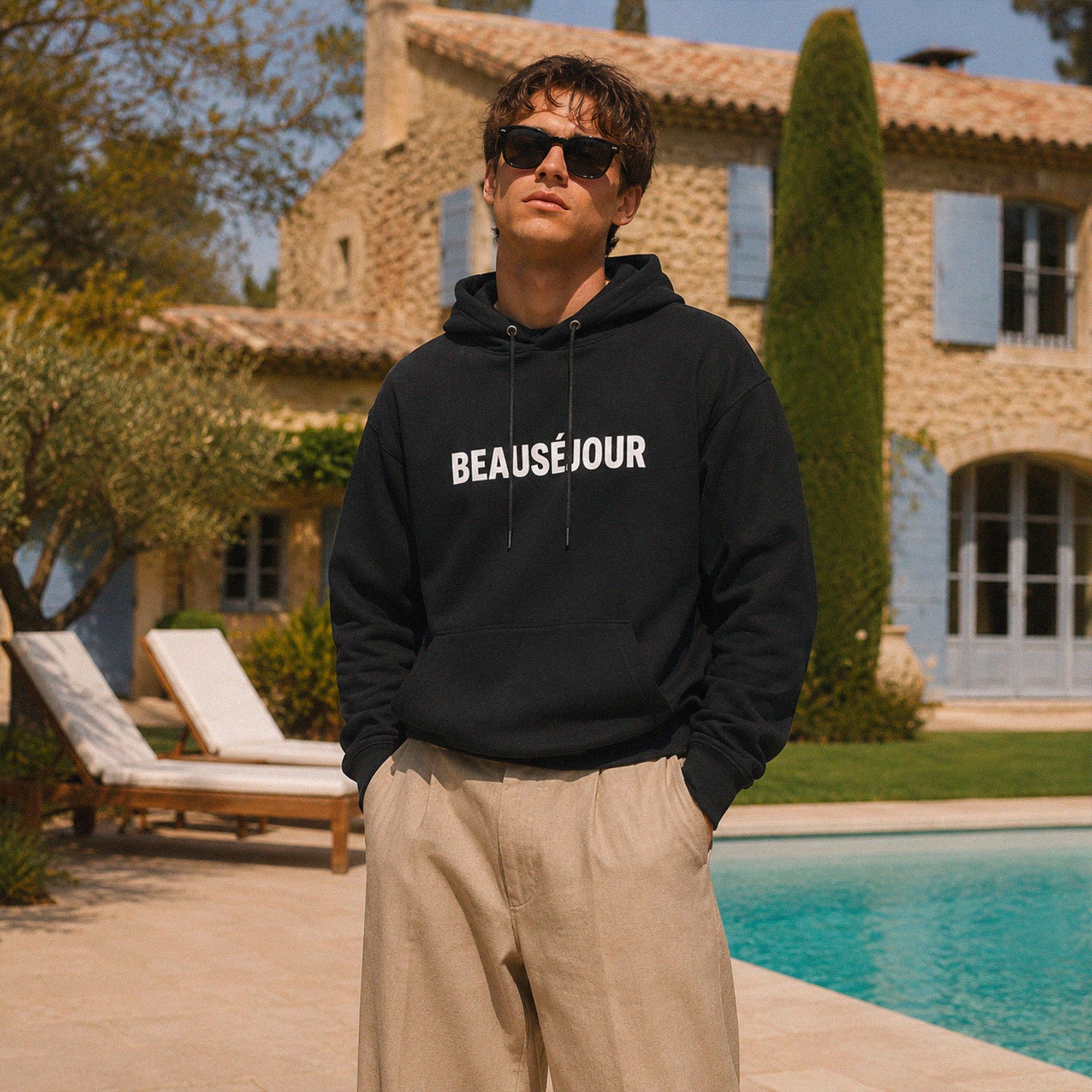 Hoodie noir Beauséjour Club de Plage avec grand imprimé palmier blanc au dos porté au bord d’une piscine dans une maison méditerranéenne.