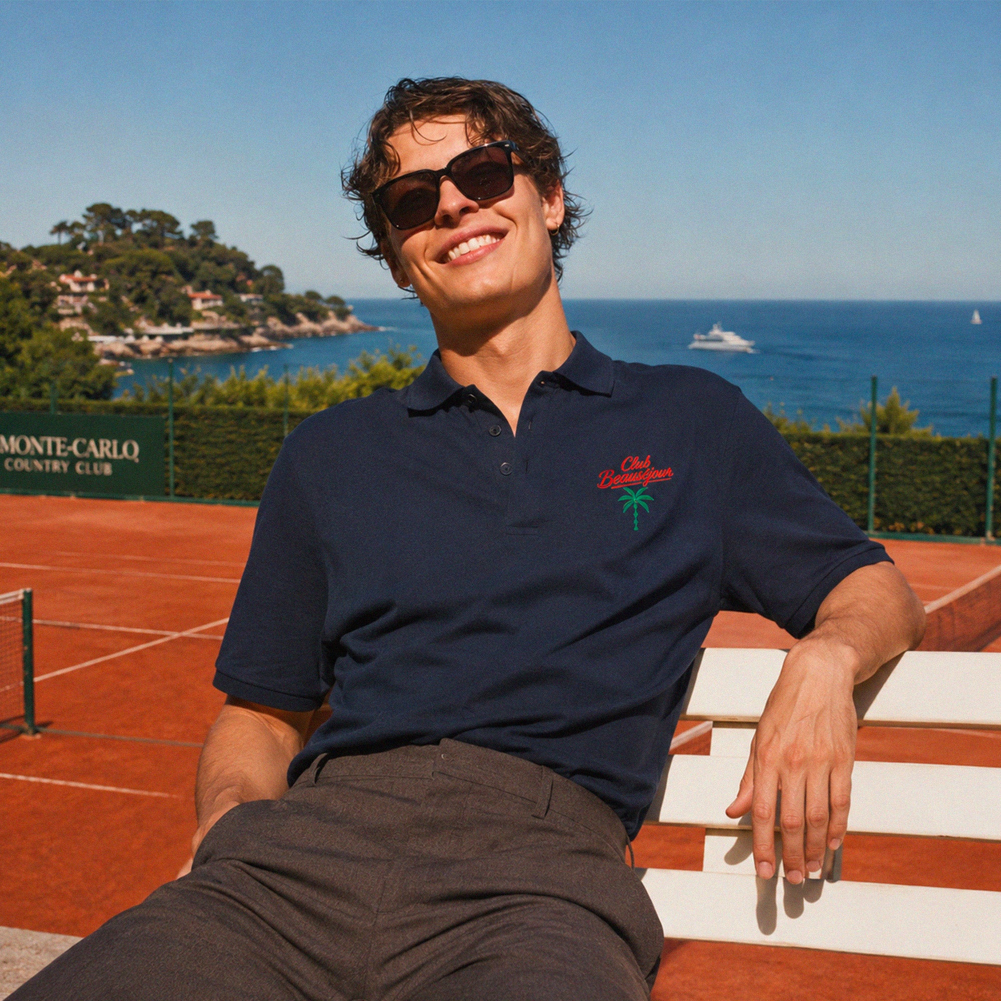 Polo bleu marine Club Beauséjour avec broderie palmier porté sur un court de tennis en bord de mer style Riviera française.