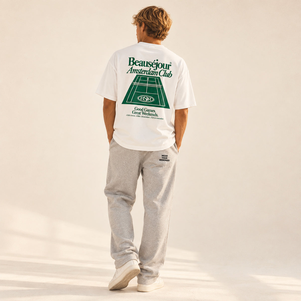 T-shirt blanc Amsterdam Club Beauséjour porté de dos par un homme, avec un grand court de tennis vert et la signature « Good Games. Great Weekends.